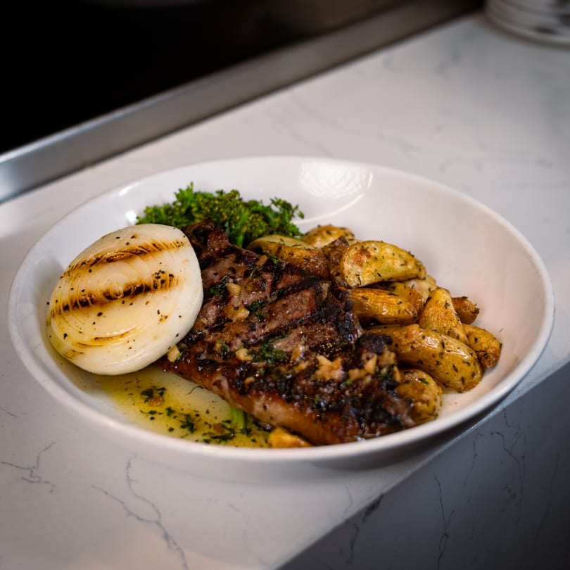 Grilled steak with roasted potatoes, charred onion, and fresh herbs in a white bowl.